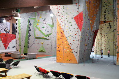 In einer Kletterhalle sind viele hohe Wände mit verschiedenen Steigungen, farbenfrohen Griffen und Kletterseilen aufgebaut.