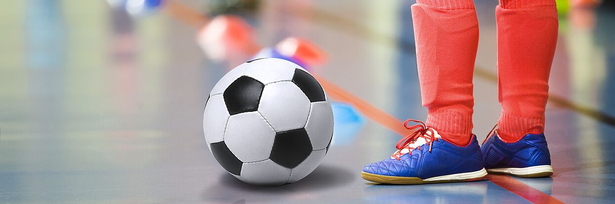 Ein Kind in Stutzen und Fußballschuhen steht vor einem Fußball in einer Indoor Soccer Halle
